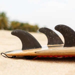 Close up view of a carbon fiber fins on a design wooden surf shortboard surfboard board at sunrise or sunset on sand beach with palm tree. Vacation concept. Summer holidays. Tourism, sport.